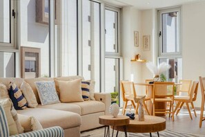 A modern living room with a beige sofa, wooden chairs, a round wooden table, and large windows. - The Reef 2C - Coral Sky (Noord)