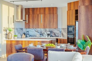 A modern kitchen with wooden cabinets, a marble backsplash, and a dining area with a table set for four. - The Reef 4A - Blue Horizon (Noord)