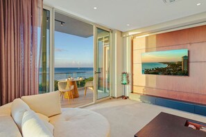 Un salon moderne avec un grand canapé, un coin repas comprenant une table et des chaises, un téléviseur fixé à un mur en bois et une porte vitrée donnant sur un balcon avec vue sur la mer.