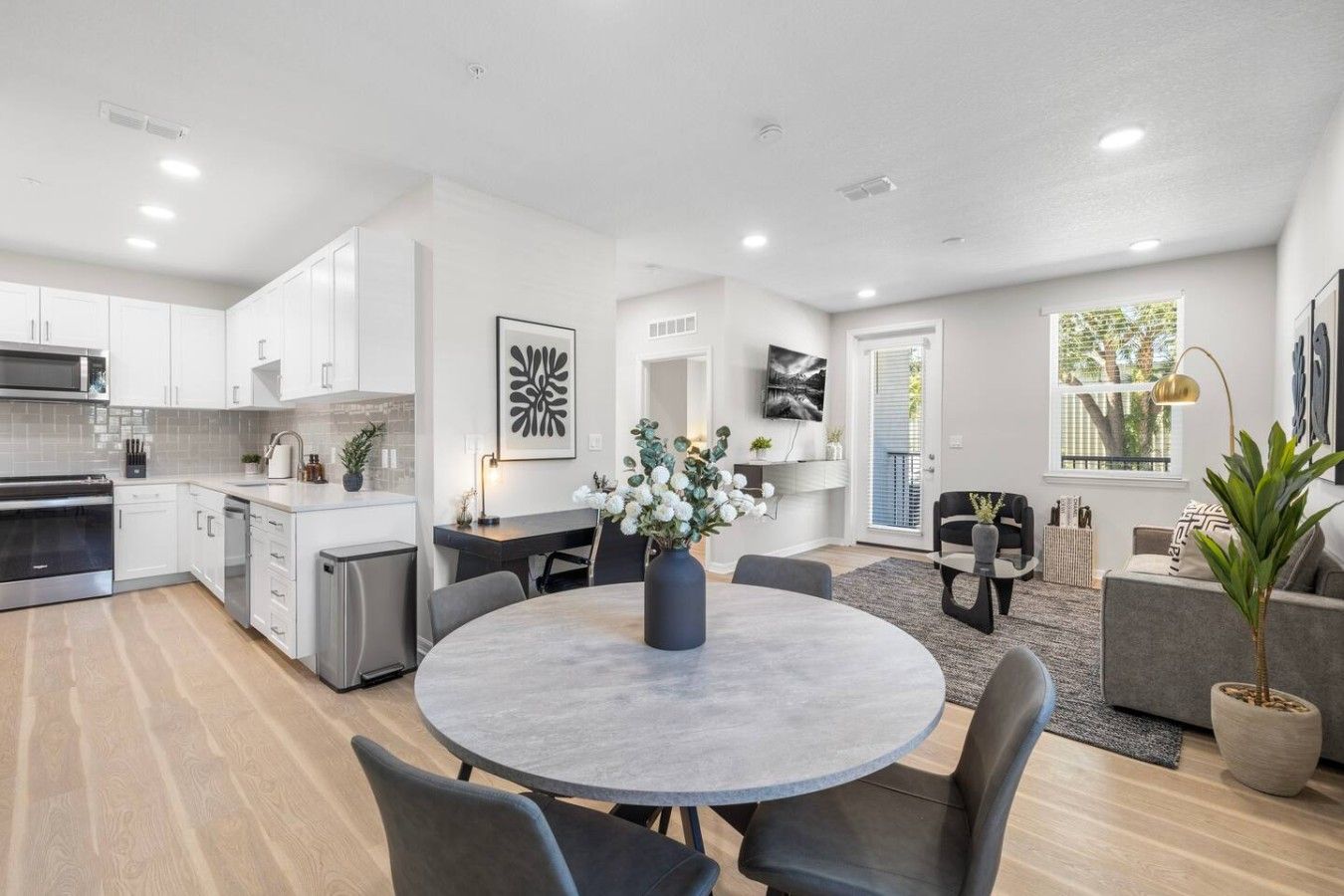 A modern kitchen with white cabinets, a dining area with a round table, and a living room with a sofa and a TV.
