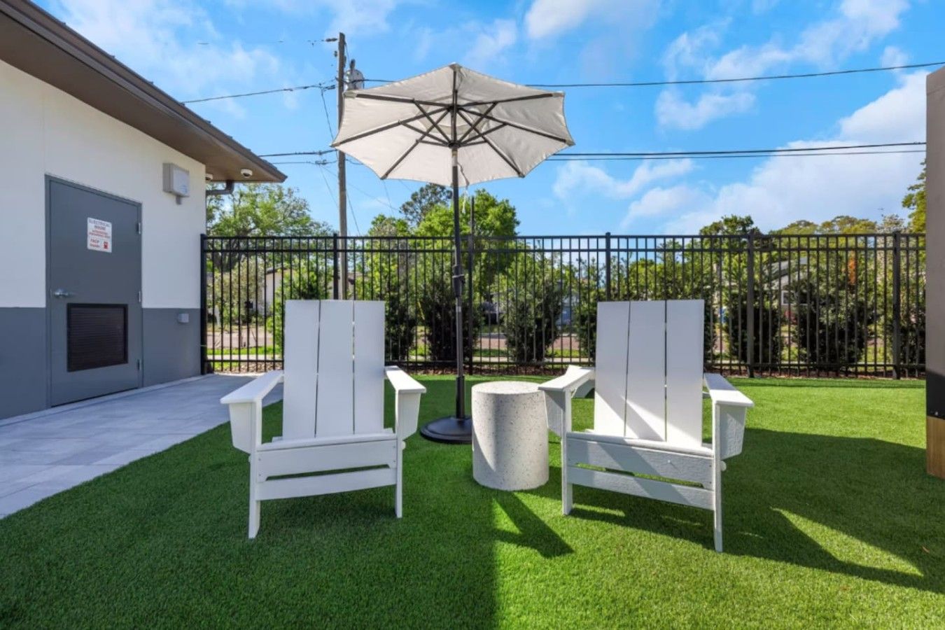 An outdoor seating area with two white chairs and a table, a beige umbrella, and a fenced yard with trees.
