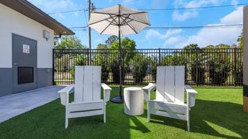 An outdoor seating area with two white chairs and a table, a beige umbrella, and a fenced yard with trees.