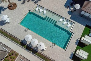 Vista aérea de una piscina con tumbonas y sombrillas, rodeada de un área pavimentada y vegetación.