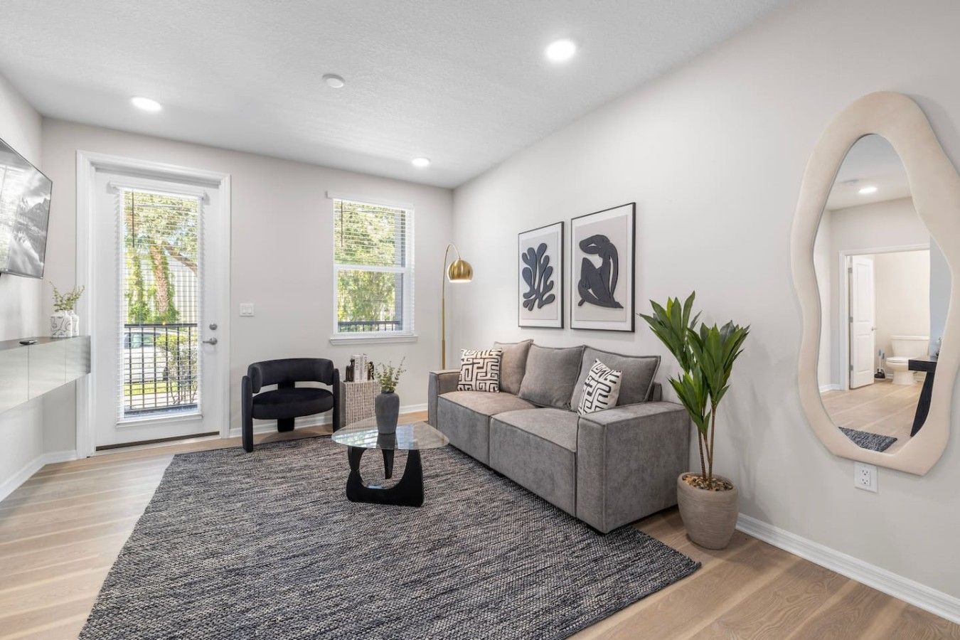 A modern living room with a grey sofa, a black armchair, a glass coffee table, and a large mirror.