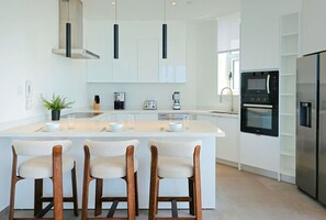 A modern kitchen with a central island, bar stools, a built-in oven, and pendant lighting. - The Reef 6A - Ocean Pearl (Noord)