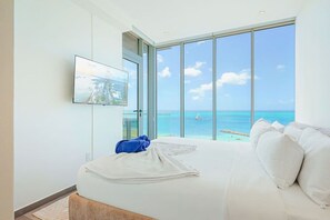 A bedroom with a large bed, a TV mounted on the wall, and a view of the ocean through large windows. - The Reef 6A - Ocean Pearl (Noord)