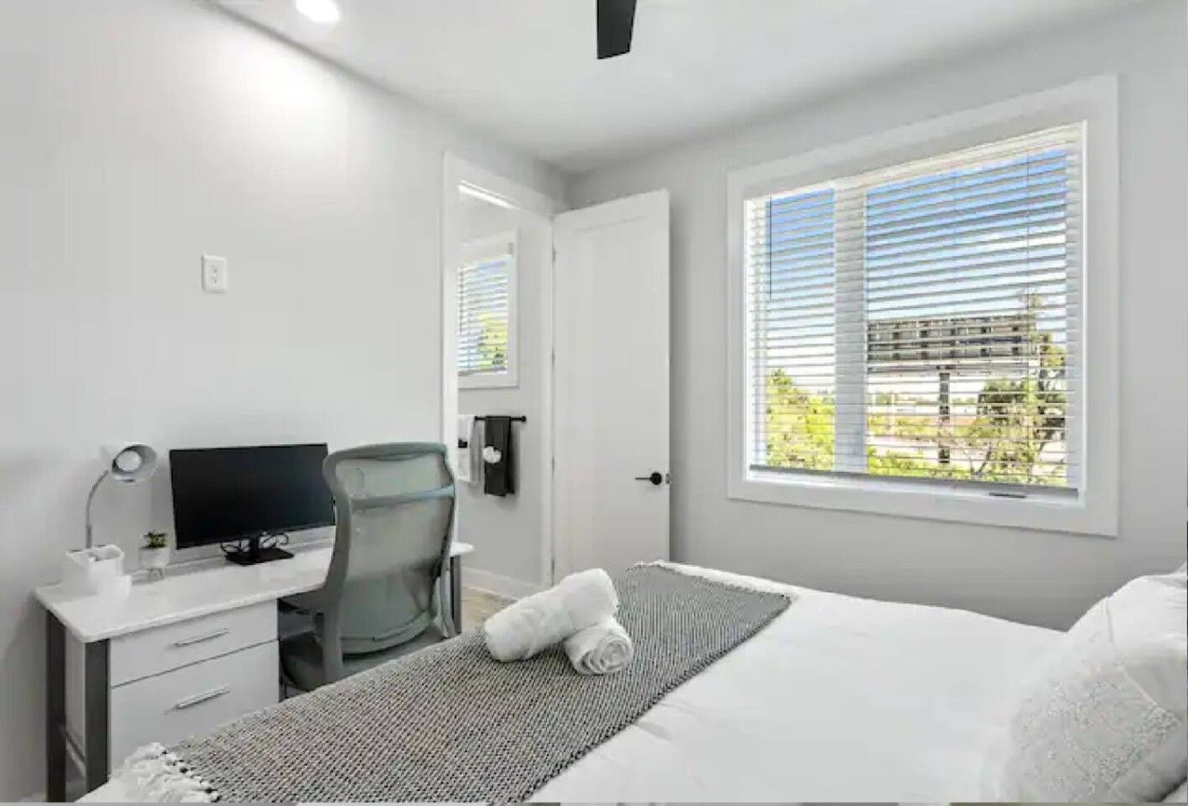 A hotel room with a bed, a desk with a computer, a chair, and a window with blinds.