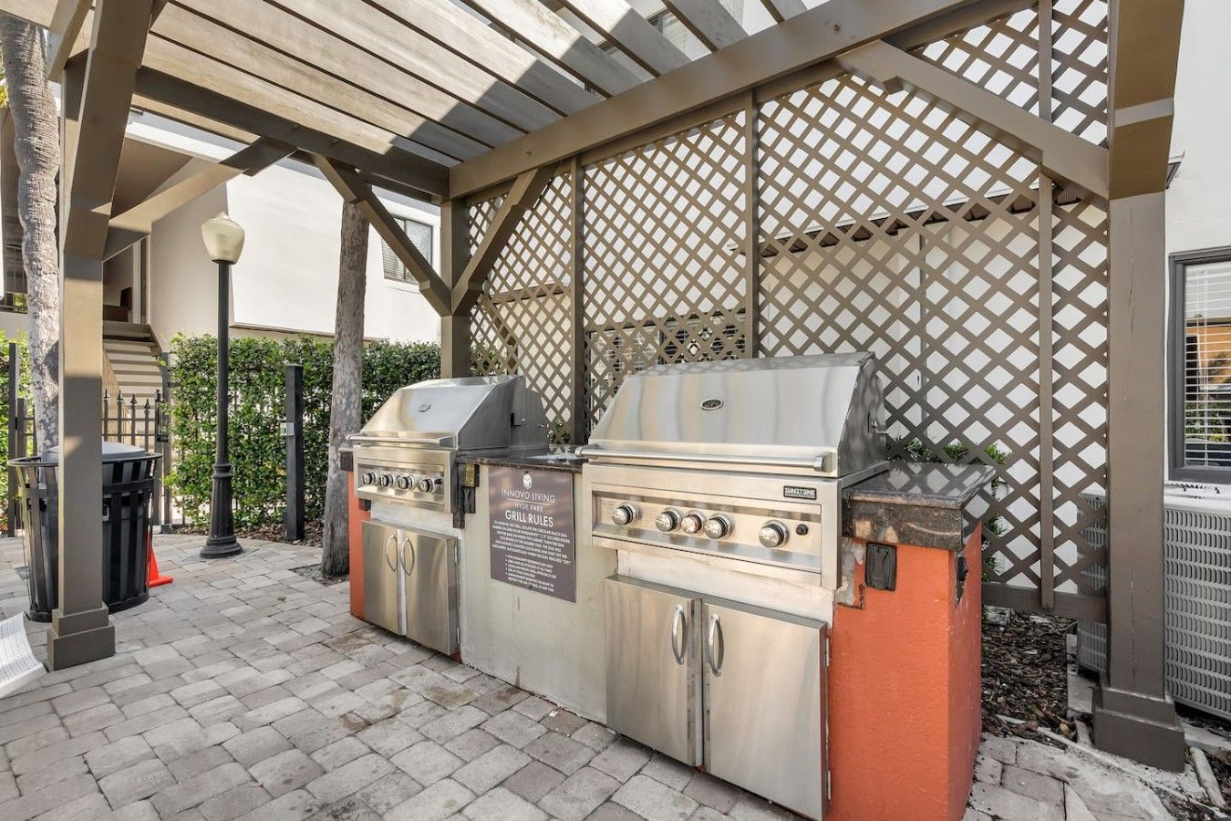 Een terras met een barbecue, een pergola erboven, een afvalbak en een straatlamp.