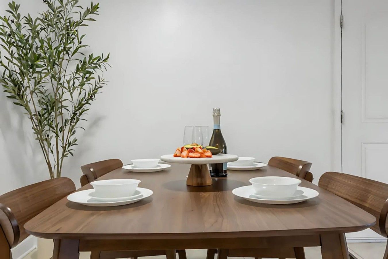 Een ronde houten eettafel met witte borden, een taartplateau in het midden en een fles wijn.