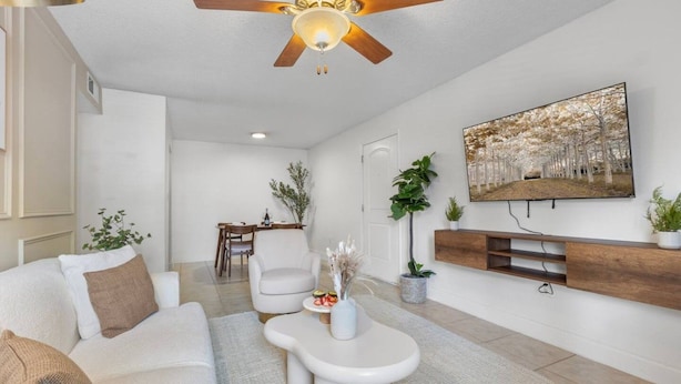 A modern living room with a white sofa, a coffee table, a ceiling fan, a dining area with a table and chairs, and a mounted TV. - Bayshore Retreat | Pool, Gym & Free Parking (Tampa)