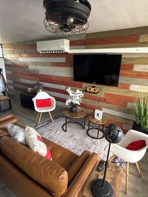 Ein modernes Wohnzimmer mit einem braunen Ledersofa, einem weißen Sessel, einem kleinen runden Tisch und einem an einer Holzwand montierten Fernseher.