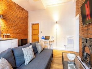 A living room with a grey sofa, a fireplace, a desk with a laptop, and a brick wall. - Student Only Ashton House (Manchester)