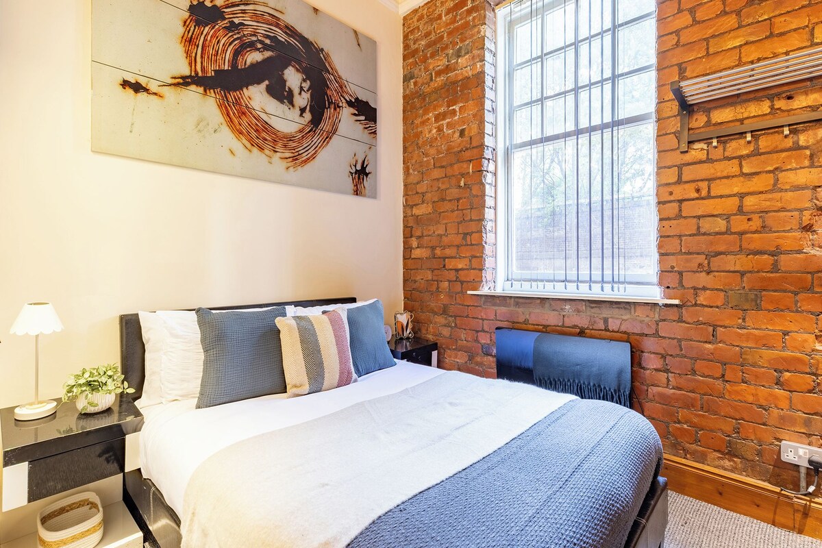 A bedroom with a brick wall, a large window, a bed with multiple pillows, a nightstand with a lamp, and a wall art piece.