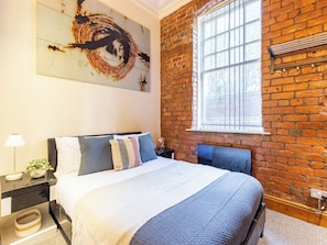 A bedroom with a brick wall, a large window, a bed with multiple pillows, a nightstand with a lamp, and a wall art piece. - Student Only Ashton House (Manchester)