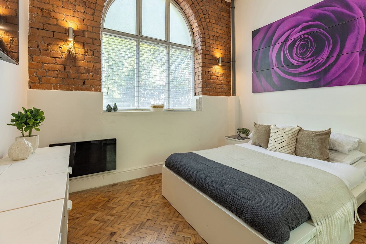 A bedroom with a brick wall, a large arched window, a bed with multiple pillows, a painting, and a small plant.
