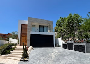 A modern two-story house with a garage, a staircase leading to the entrance, and a landscaped garden with cacti and shrubs. - Villa Velas | Private Pool, Hot Tub & Rooftop | Gated Cabo Bello (Cabo San Lucas)