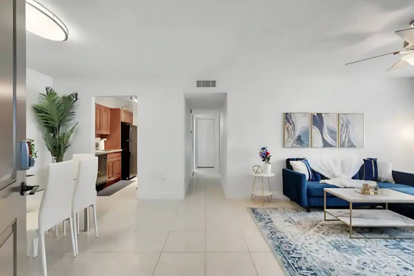 Ein modernes Wohnzimmer mit einem blauen Sofa, einem weißen Couchtisch und einem kleinen Beistelltisch mit einer Vase. Im Hintergrund ist eine Küche mit Holzschränken und einem Kühlschrank zu sehen.