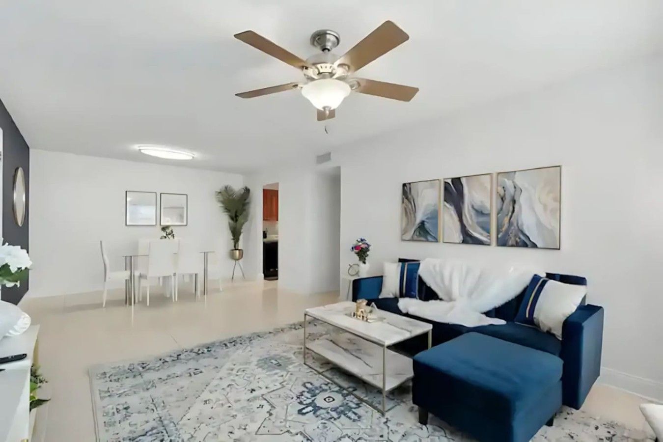 Uma sala de estar moderna com um ventilador de teto, um sofá azul, uma mesa de centro e uma área de jantar com mesa e cadeiras brancas.