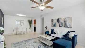 Uma sala de estar moderna com um ventilador de teto, um sofá azul, uma mesa de centro e uma área de jantar com mesa e cadeiras brancas.