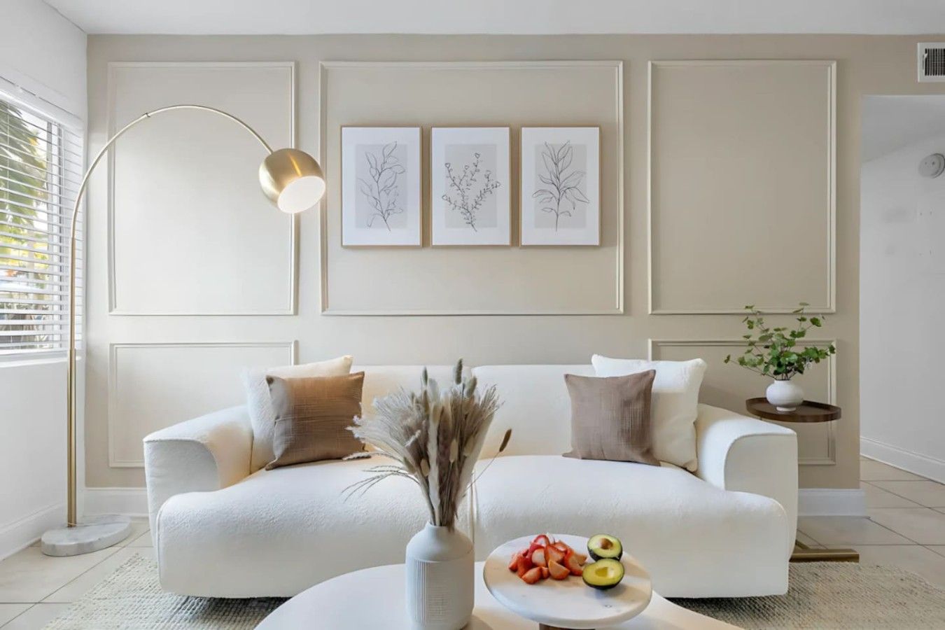 A modern living room with a white sofa, decorative pillows, a floor lamp, and framed botanical prints on the wall.