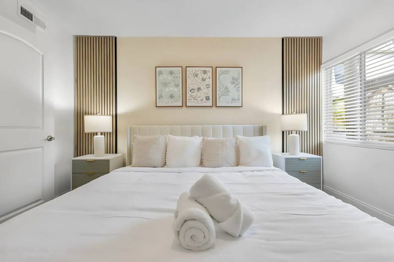 Une chambre d’hôtel avec un grand lit, deux tables de chevet, une tête de lit et trois tableaux encadrés accrochés au mur.