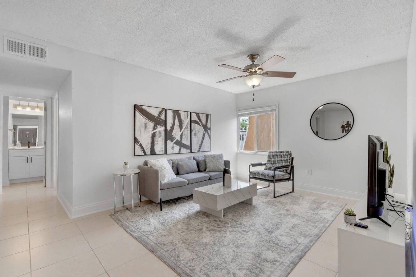 Un salon moderne avec un canapé gris, une table basse, un ventilateur de plafond, un téléviseur à écran plat et un grand tableau abstrait.