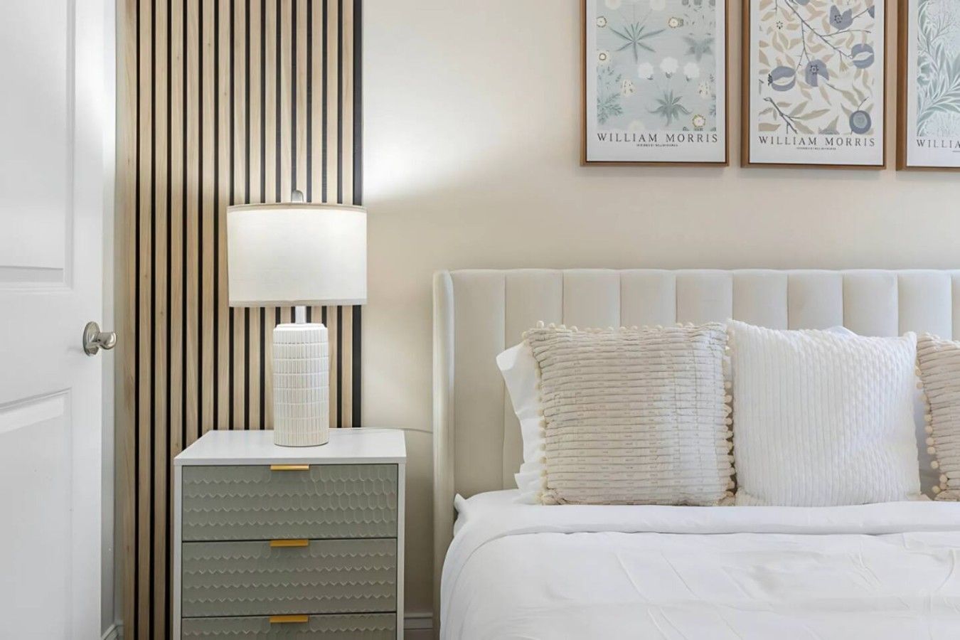 A bedroom with a white bed, two pillows, a bedside table with a lamp, and a wall with framed artwork.