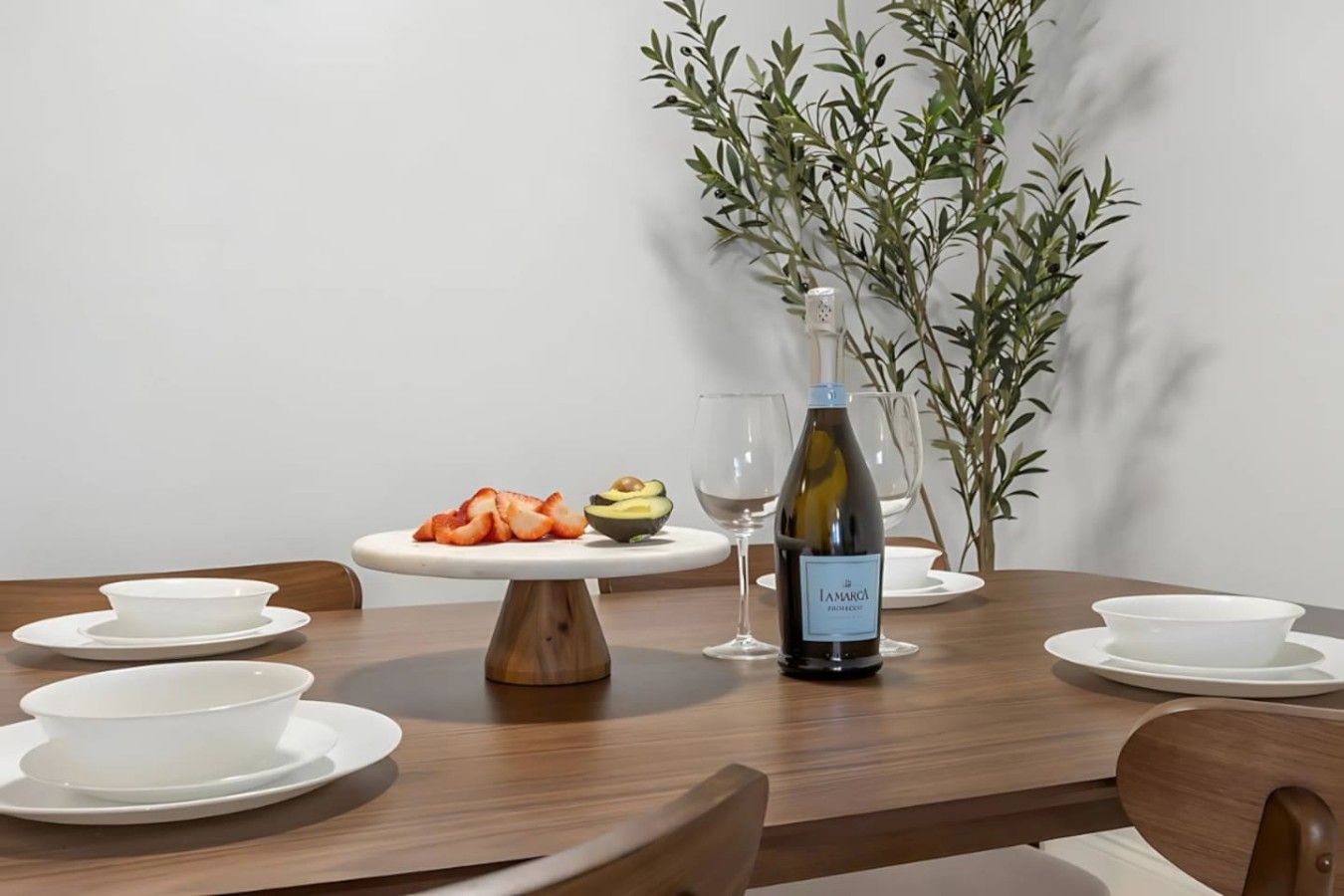 A dining table set with white plates, a white bowl, a bottle of wine, and a plate of sliced fruits.