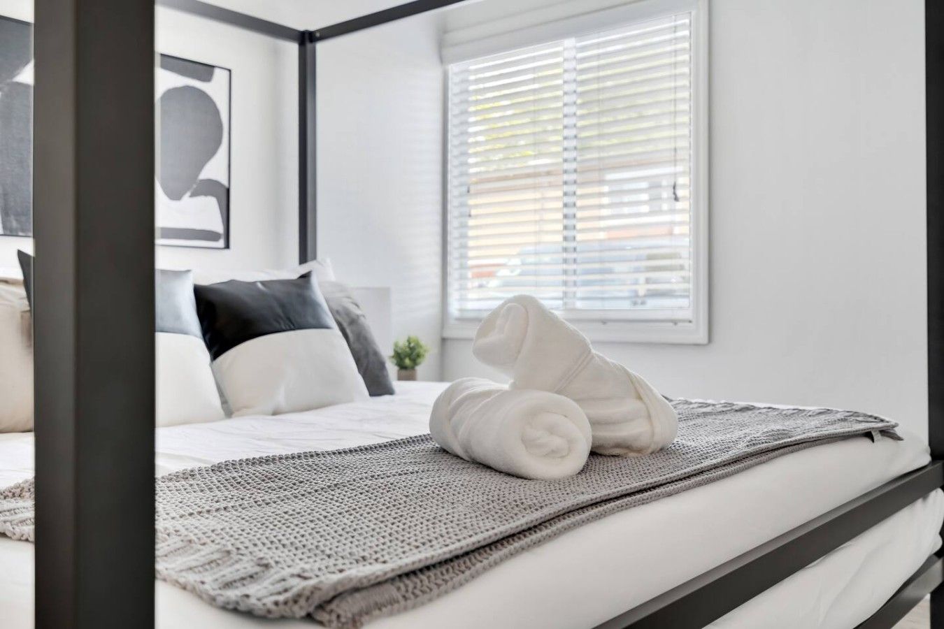Cama bem arrumada com edredão branco e toalhas enroladas brancas, um cobertor de tricô cinza, e uma janela com persianas.