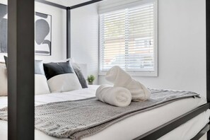 Cama bem arrumada com edredão branco e toalhas enroladas brancas, um cobertor de tricô cinza, e uma janela com persianas.