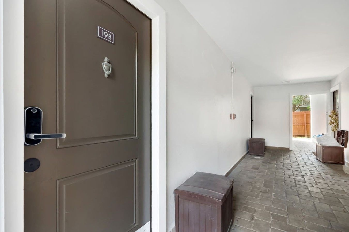 A hallway with a brown door marked '19B', a small bench, and a view of a wooden fence outside.
