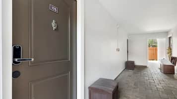 A hallway with a brown door marked '19B', a small bench, and a view of a wooden fence outside.