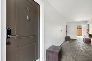 Corredor com uma porta marrom com a inscrição '19B', um pequeno banco e vista para uma cerca de madeira.