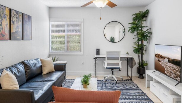 A modern living room with a leather sofa, a desk with a chair, a ceiling fan, a large mirror, and a television. - South Tampa Coastal Stay | Pool & Free Parking (Tampa)