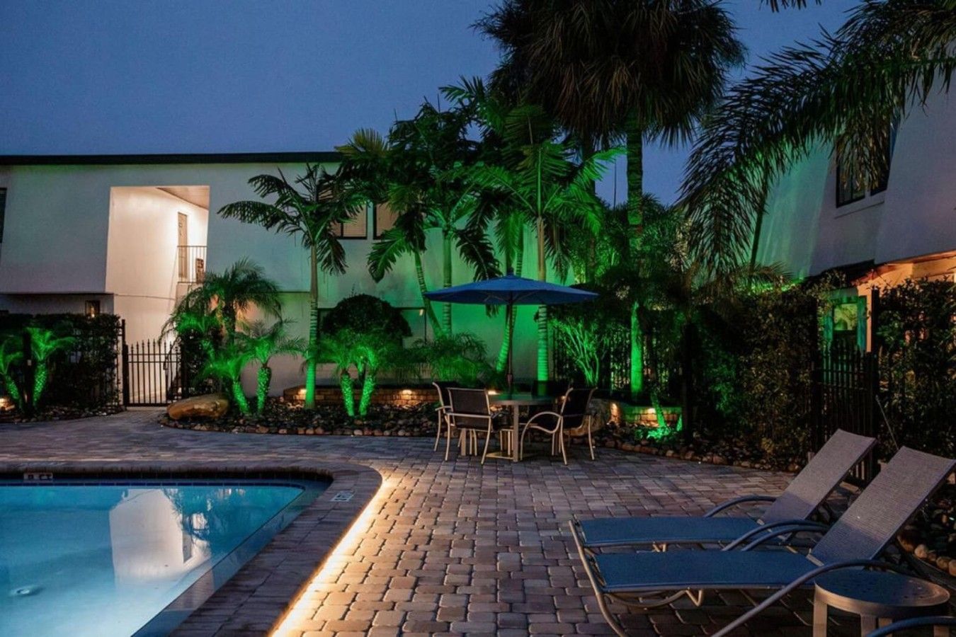 A swimming pool with lounge chairs and a table, surrounded by palm trees and a building with a modern design.