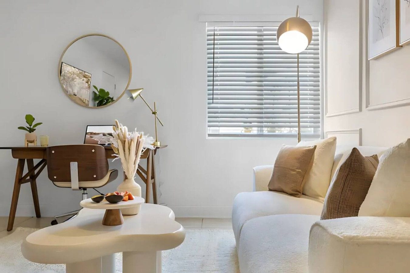 A modern living room with a sofa, a round mirror, a desk with a chair, and a window with blinds.