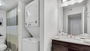 A bathroom with a white toilet, a washing machine, a sink with a mirror, and a shower curtain. - Palma Ceia Retreat | Pool, Gym & Free Parking (Tampa)