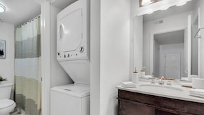 A bathroom with a white toilet, a washing machine, a sink with a mirror, and a shower curtain. - Bayshore Bliss | Pool, Gym & Free Parking (Tampa)