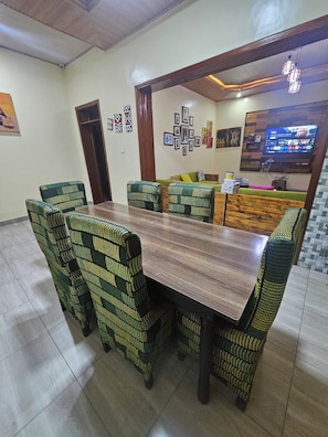 Een eetgedeelte met een houten tafel en gestreepte stoelen, een groene bank en een aan de muur bevestigde televisie.