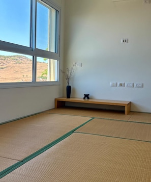 A room with tatami flooring, a window with a view of a dry landscape, a wooden bench, a vase with dried plants, and a wall with electrical outlets.