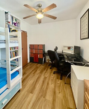 Una stanza con pavimento in legno, una scrivania con computer, una sedia, una libreria e una ventola a soffitto.