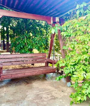 A wooden swing under a covered area with greenery around.