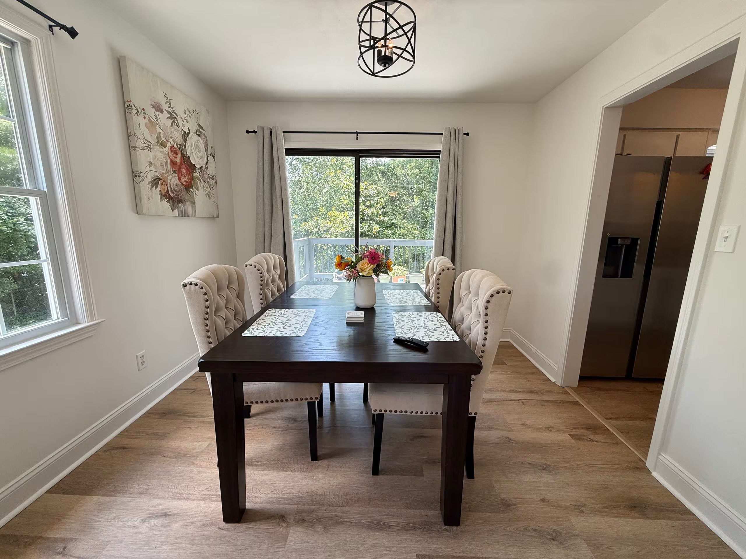 Een eetgedeelte met een donker houten tafel, zes gestoffeerde stoelen, een vaas met bloemen en een uitzicht door een groot raam.