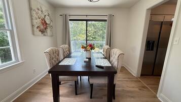Een eetgedeelte met een donker houten tafel, zes gestoffeerde stoelen, een vaas met bloemen en een uitzicht door een groot raam.