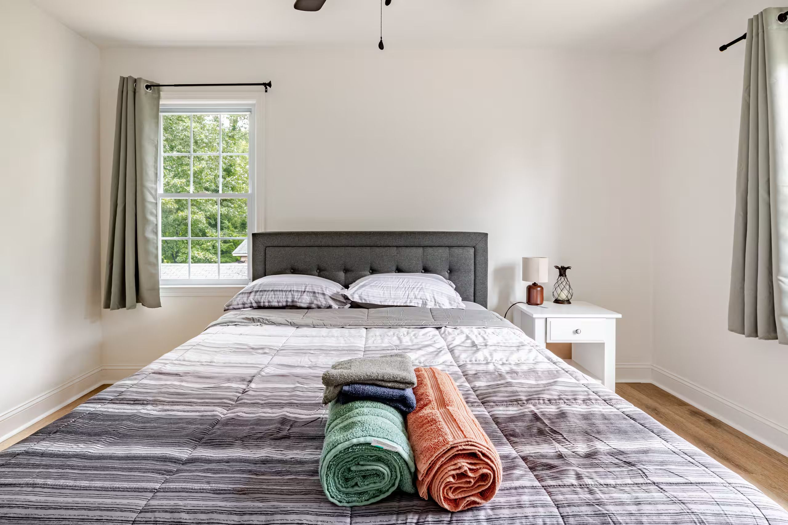 Een slaapkamer met een groot bed, een nachtkastje, een raam met gordijnen en uitzicht op bomen.