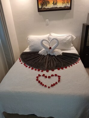 A bed with a white bedspread, a dark brown skirt, and a heart-shaped decoration made of rose petals.