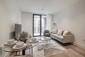 A modern living room with a sofa, a glass coffee table, and a dining area with plates and glasses. - Elegant One-Bedroom Retreat (London)