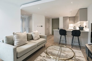 A modern living room with a sofa, two chairs, a glass coffee table, and a kitchen area with a dining table. - Stylish Two-Bedroom Residence (London)