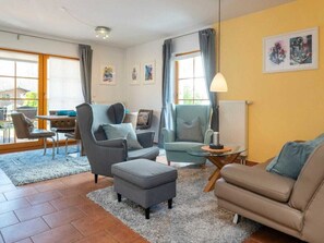 A living room with a sofa, armchair, ottoman, and a small table with a lamp. There are two windows with curtains, a radiator, and artwork on the wall. - 7b/t5 Holiday Home Park \"freesenbruch\" (Zingst)