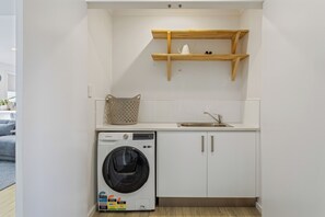 A compact laundry room with a washing machine, a sink, and a wooden shelf above. - Invermay Escape Stay Close to Stadium and Museums (Invermay)